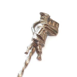 May include: A silver-toned metal spoon with a decorative figure of a person carrying a basket on their back. The spoon's handle has a twisted design.