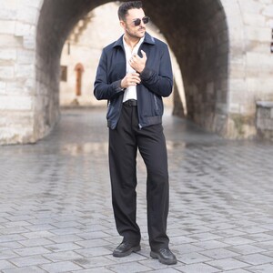 Puede incluir: Un hombre con una chaqueta azul oscuro con una camisa blanca debajo, pantalones negros y zapatos negros. Está de pie frente a un arco.