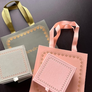 May include: Two gift bags and two jewellery boxes. One bag is sage green with a gold ribbon handle, and the other is pink with a pink ribbon handle. The boxes are sage green and pink, with gold trim and a white ribbon pull.