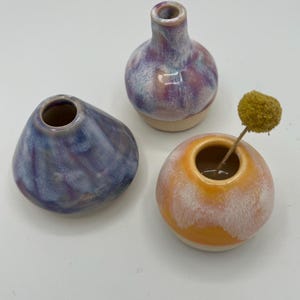 Può includere: Tre piccoli vasi in ceramica nei toni del blu, viola e arancione. Uno ha la forma di una goccia arrotondata, un altro è di forma bulbosa e il terzo è rotondo con un fiore secco. I vasi hanno una finitura lucida.