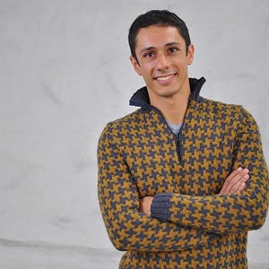 May include: A patterned sweater in shades of mustard yellow and navy blue. The sweater has a zipper at the collar and a dark blue collar. The person is smiling with arms crossed.