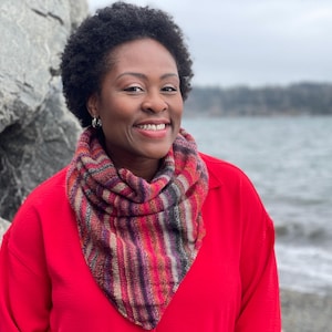 May include: A triangular knit scarf featuring a variegated pattern of magenta, purple, orange, and dark grey stripes. The scarf is draped around the neck, showcasing a textured, hand-knitted stitch pattern with a pointed front edge.