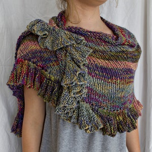 May include: A multi-colored knitted shawl with a ruffled edge. The shawl is made with a variety of colors, including purple, pink, green, and gold. The shawl is draped over a person's shoulders.