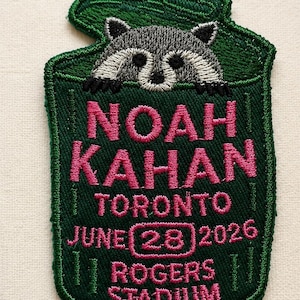 Op de afbeelding: Een donkergroene geborduurde patch in de vorm van een vuilnisbak met een wasbeer die bovenuit steekt. De tekst NOAH KAHAN TORONTO JUNE 28 2026 ROGERS STADIUM is in roze draad geborduurd. De wasbeer is gestikt in tinten grijs, zwart en wit.