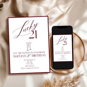 May include: A white invitation with burgundy borders and text that reads "Lucky 21" and "You are invited to celebrate Amelia's 21st birthday." A smartphone displays the same invitation. The invitation is on a gold tray.