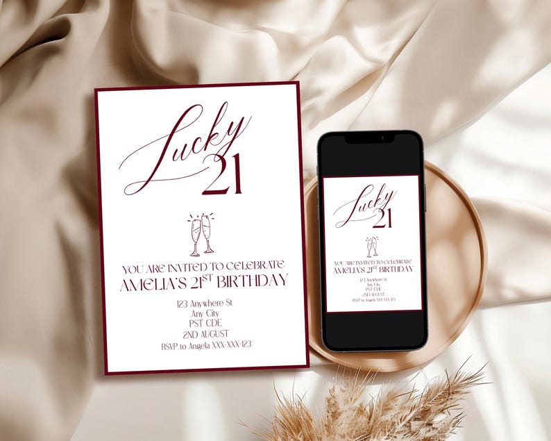 May include: A white invitation with burgundy borders and text that reads "Lucky 21" and "You are invited to celebrate Amelia's 21st birthday." A smartphone displays the same invitation. The invitation is on a gold tray.