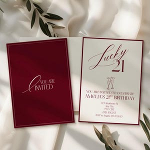 May include: Burgundy and cream-colored invitation cards. One card reads "You are Invited" in white script. The other card says "Lucky 21" with event details. The cards are on a white background with greenery.