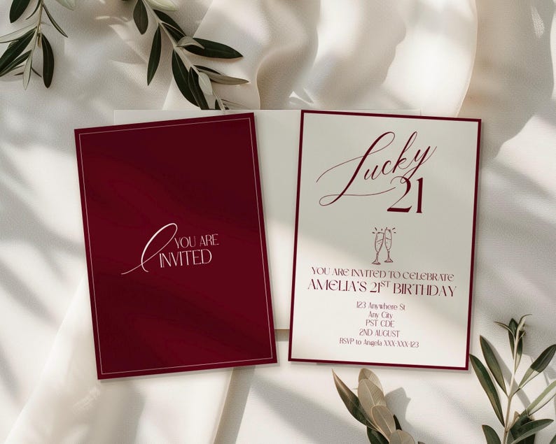 May include: Burgundy and cream-colored invitation cards. One card reads "You are Invited" in white script. The other card says "Lucky 21" with event details. The cards are on a white background with greenery.