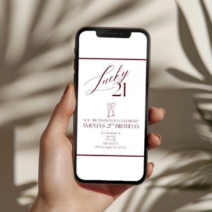 May include: A smartphone displays a digital invitation for a "Lucky 21" birthday celebration. The invitation features elegant burgundy text on a white background, including the name "Amelia's" and event details. The phone is held in a person's hand.