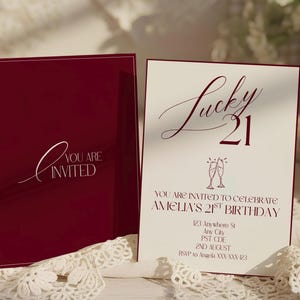May include: Burgundy invitation card with the text "You are Invited" in white script. The open card displays "Lucky 21" in burgundy script, with details for Amelia's 21st birthday, including date, time, and RSVP information.