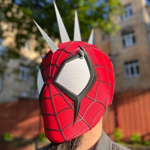May include: A red and black Spider-Man mask with white eye lenses and silver spikes on top. The mask has a web pattern and black trim. The mask is a cosplay item.