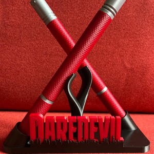 Könnte beinhalten: Ein rot-silberner Stifthalter mit zwei gekreuzten Stiften. Die Stifte haben ein strukturiertes Muster und silberne Akzente. Die schwarze Basis zeigt das Wort "DAREDEVIL" in Rot mit einer Stadtsilhouette.