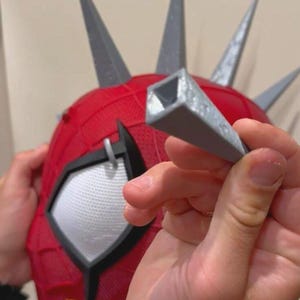 May include: Red and gray superhero mask with gray spikes. The mask has white mesh eye coverings and a black outline. A gray, square-shaped piece is held in front of the mask.
