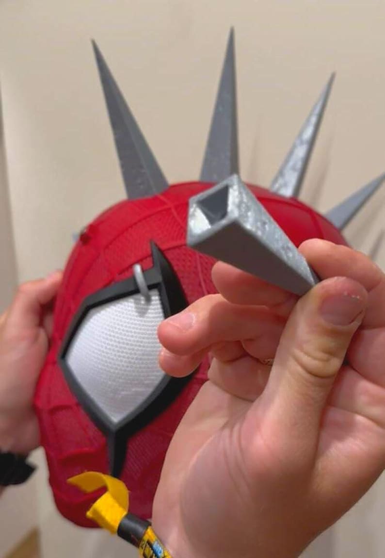 May include: Red and gray superhero mask with gray spikes. The mask has white mesh eye coverings and a black outline. A gray, square-shaped piece is held in front of the mask.