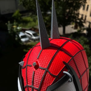 May include: A red and black Spider-Man mask with a web pattern, gray spikes, and a mesh eye covering. The mask has a textured surface and is designed to resemble the superhero's iconic look.