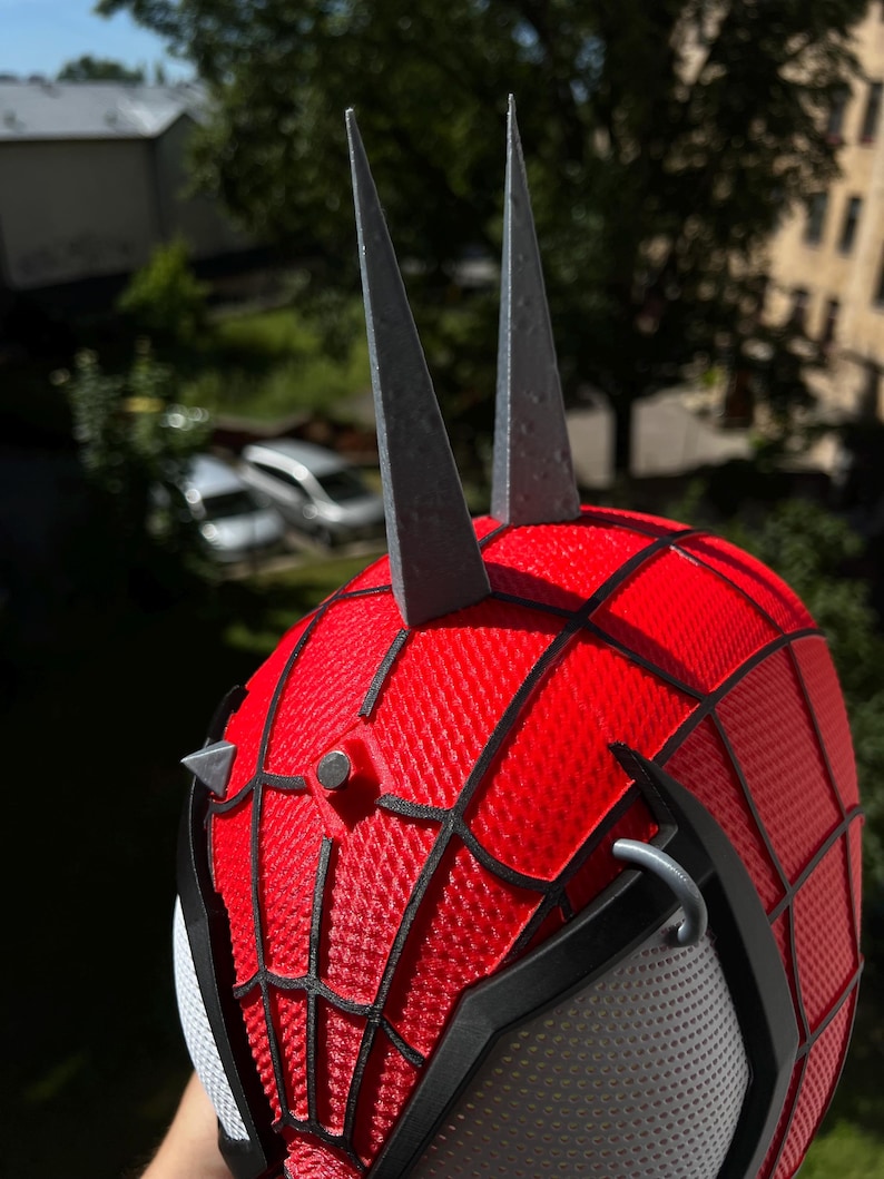 May include: A red and black Spider-Man mask with a web pattern, gray spikes, and a mesh eye covering. The mask has a textured surface and is designed to resemble the superhero's iconic look.