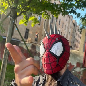May include: A red and black Spider-Man helmet with white eye lenses and silver spikes. The helmet has a webbed design and is worn outdoors with a blurred background of trees and buildings.