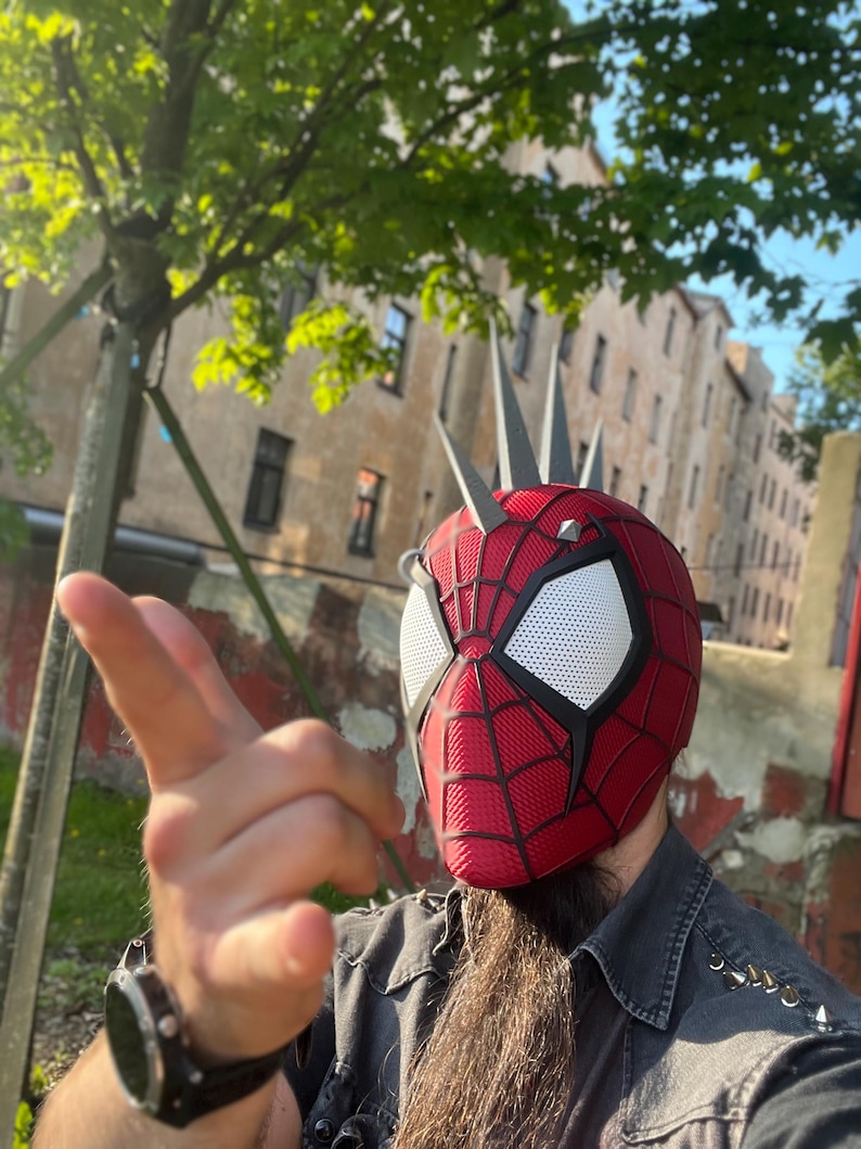 May include: A red and black Spider-Man helmet with white eye lenses and silver spikes. The helmet has a webbed design and is worn outdoors with a blurred background of trees and buildings.
