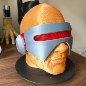 Op de afbeelding: Een 3D-geprinte oranje buste met een grijze visor met een rood mesh detail. De buste staat op een zwarte cirkelvormige basis, met een Batman-vormig object op de achtergrond. De visor heeft een futuristisch ontwerp.