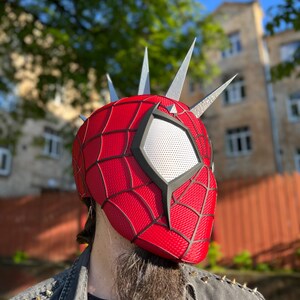 May include: A red and black Spider-Man mask with silver spikes. The mask has a webbed pattern and a white eye section with a mesh covering. The mask is worn with a black shirt and a denim vest with silver spikes.