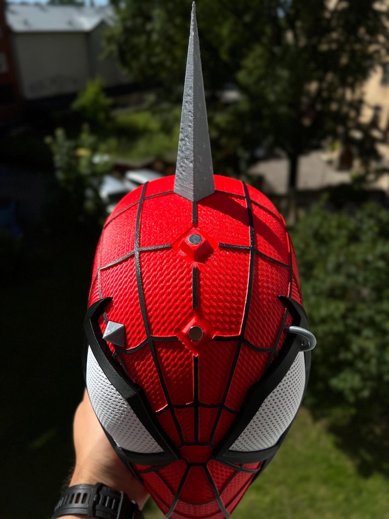 May include: A red and black Spider-Man mask with a silver spike on top. The mask has a web pattern and white mesh eye coverings. The mask is held up against a green background.