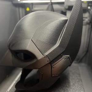 Könnte beinhalten: Eine schwarze Batman-Maske mit strukturierter Oberfläche und spitzen Ohren. Die Maske hat ein schlankes, modernes Design mit detaillierten Paneelen und Nieten. Die Augenöffnungen sind sichtbar, und die Gesamtästhetik ist düster und imposant.