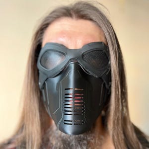 May include: A black, futuristic-style mask with mesh eye coverings and a vented lower section. The mask is made of a matte material and has a detailed, angular design. The mask is worn by a person with long brown hair and a beard.