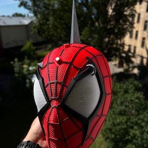May include: A red and black Spider-Man mask with white mesh eye coverings and a silver spike on top. The mask has a textured pattern and is held up against a background of green trees and a blue sky.