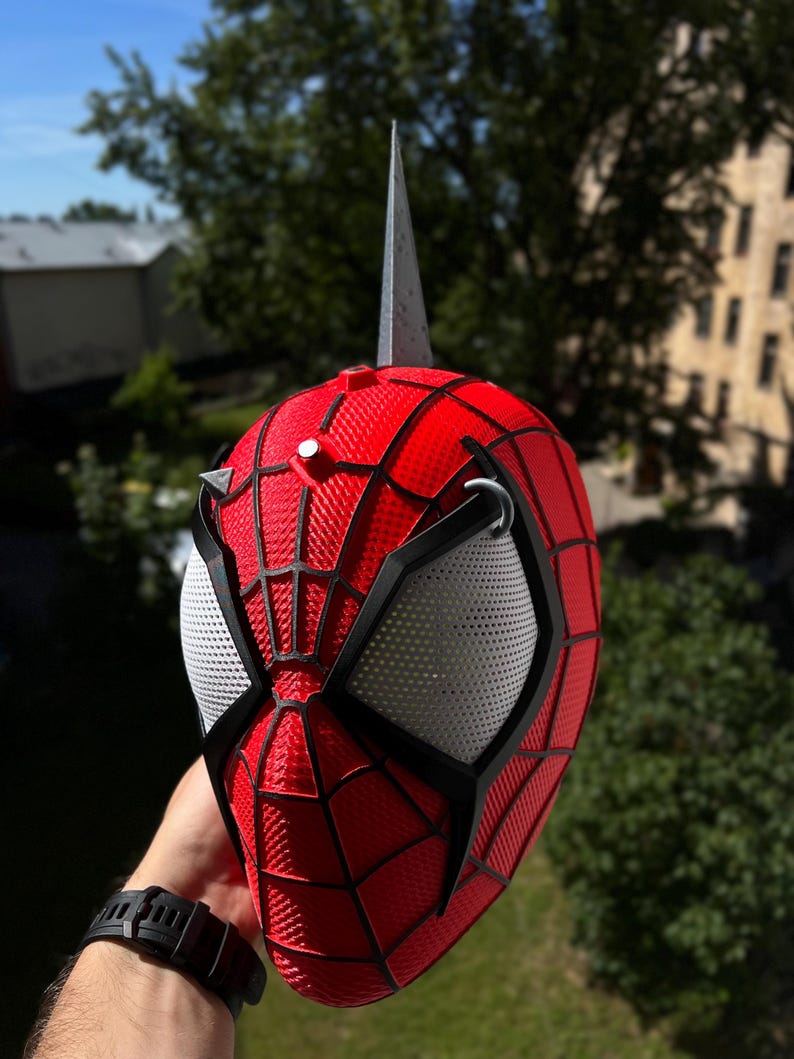 May include: A red and black Spider-Man mask with white mesh eye coverings and a silver spike on top. The mask has a textured pattern and is held up against a background of green trees and a blue sky.