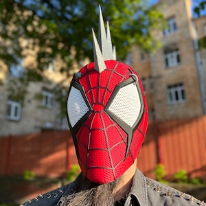 May include: A red and black Spider-Man mask with white eye lenses and silver spikes on top. The mask has a webbed design and is worn by a person with a long beard and a denim jacket.