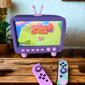 May include: A purple retro TV with a screen displaying the Animal Crossing: New Horizons game. Two pink and blue Nintendo Switch Joy-Con controllers are on a wooden surface.