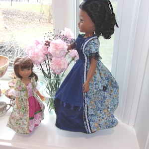 Könnte beinhalten: Zwei Puppen werden auf einer weißen Fensterbank ausgestellt. Eine Puppe trägt ein blaues Kleid mit Blumenmustern und einen dunkelblauen Rock. Die andere Puppe ist kleiner und trägt ein rosa Kleid mit einem Mantel mit Blumenmuster. Rosa Blumen sind in einer Vase.