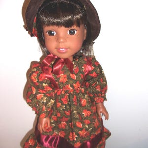 Könnte beinhalten: Puppe mit braunen Haaren, Sommersprossen und braunen Augen, die einen braunen Hut und ein Kleid mit einem Herbstblattmuster in Rot, Orange und Grün trägt. Das Kleid hat ein bordeauxfarbenes Band um die Taille und den Halsausschnitt.
