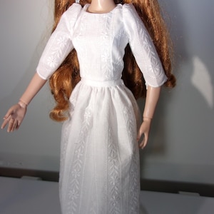 May include: A doll in a white dress featuring floral embroidery. The dress has three-quarter sleeves and a fitted bodice that flows into a flared skirt. The doll has long, red hair and is wearing earrings.