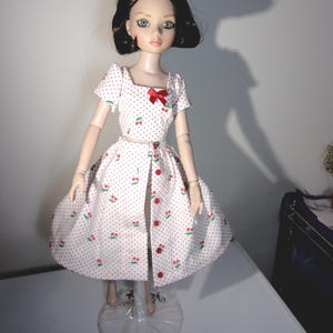 Könnte beinhalten: Puppe mit dunklem Haar und blauen Augen, die ein weißes Kleid mit rotem Kirsch- und Tupfenmuster trägt. Das Kleid hat eine rote Schleife und rote Knöpfe. Die Puppe steht auf einem transparenten Ständer.