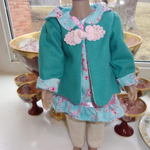 May include: A doll in a teal jacket with pink floral details and a matching floral dress. The jacket features pink decorative fastenings. The doll is wearing beige trousers. The outfit is displayed with decorative bowls and a teacup.