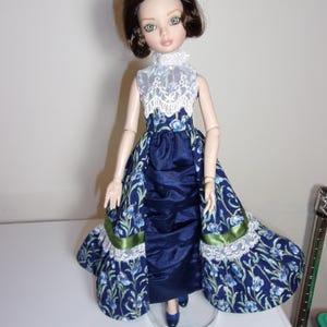 May include: A doll wearing a navy blue dress with a floral pattern and a white lace collar. The dress has a satin skirt and a floral overskirt with green ribbon trim. The doll has brown hair and blue shoes.