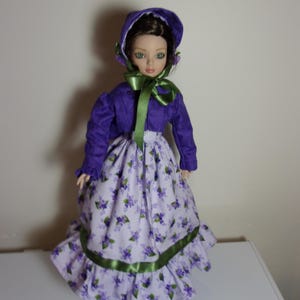 Peut inclure: Une poupée vêtue d'une tenue violette, comprenant un bonnet et une longue jupe à motifs floraux et garniture de ruban vert. La veste de la poupée est ornée d'un nœud en ruban vert. La poupée a les yeux verts et les cheveux bruns.
