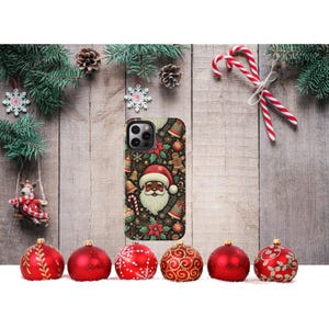 May include: A phone case with a festive Christmas design featuring Santa Claus, gingerbread men, and candy canes. The case is surrounded by red ornaments, pine branches, and snowflakes. The background is a wooden surface.