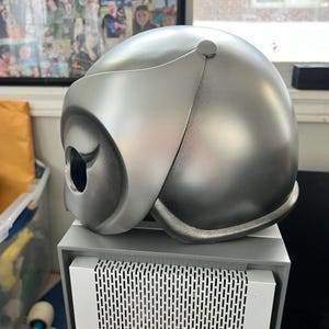 May include: A silver helmet with a visor and a black owl design. The helmet is made of a smooth, hard material and has a matte finish.
