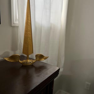 Puede incluir: Una escultura de metal dorado de una lanza con alas. La lanza tiene una punta afilada y las alas están extendidas a ambos lados. La escultura está sobre una superficie de madera oscura. La escultura tiene la palabra "EXCELLENCE" inscrita en la base.