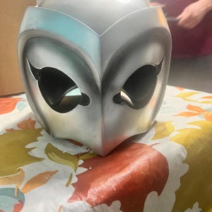 May include: A silver-colored mask with a stylized owl design. The mask has two large eye holes and a pointed beak.