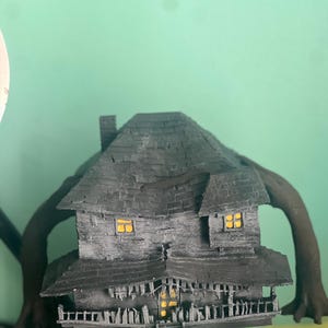May include: A black 3D printed model of a house with a crooked roof and four spindly arms. The house has windows with yellow lights and a porch with a railing. The model is sitting on a green jar.