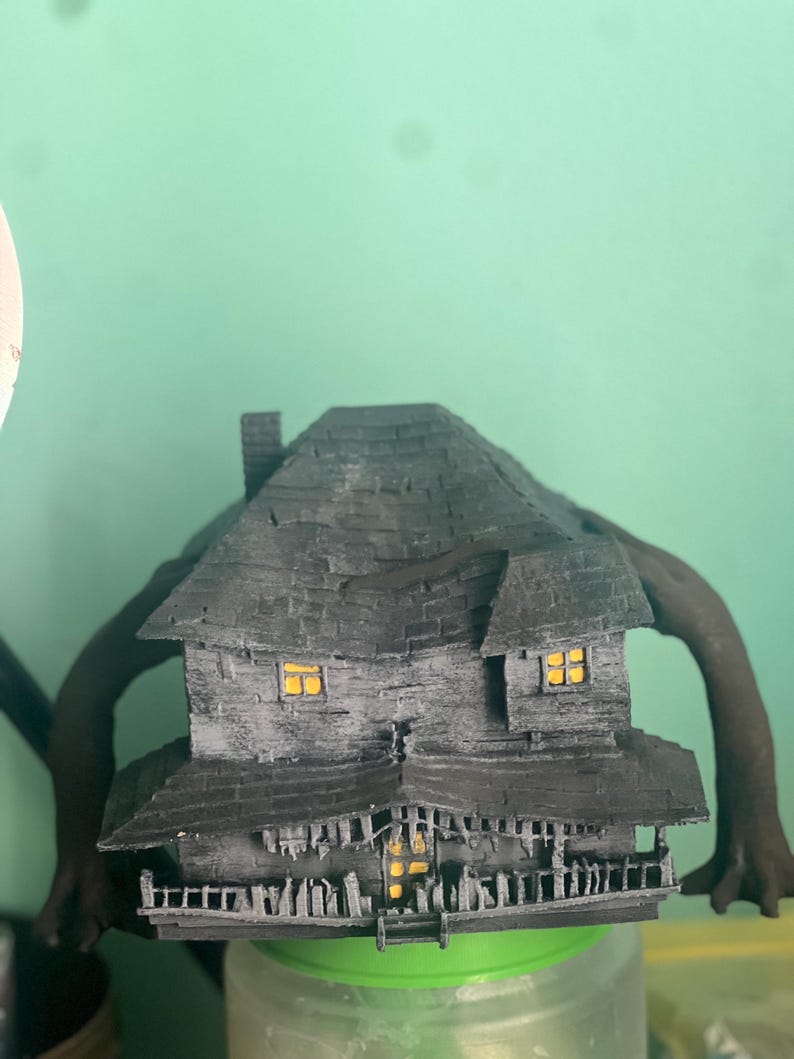 May include: A black 3D printed model of a house with a crooked roof and four spindly arms. The house has windows with yellow lights and a porch with a railing. The model is sitting on a green jar.