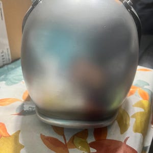 May include: A silver helmet with a smooth, shiny surface. The helmet has two metal clasps on the side.