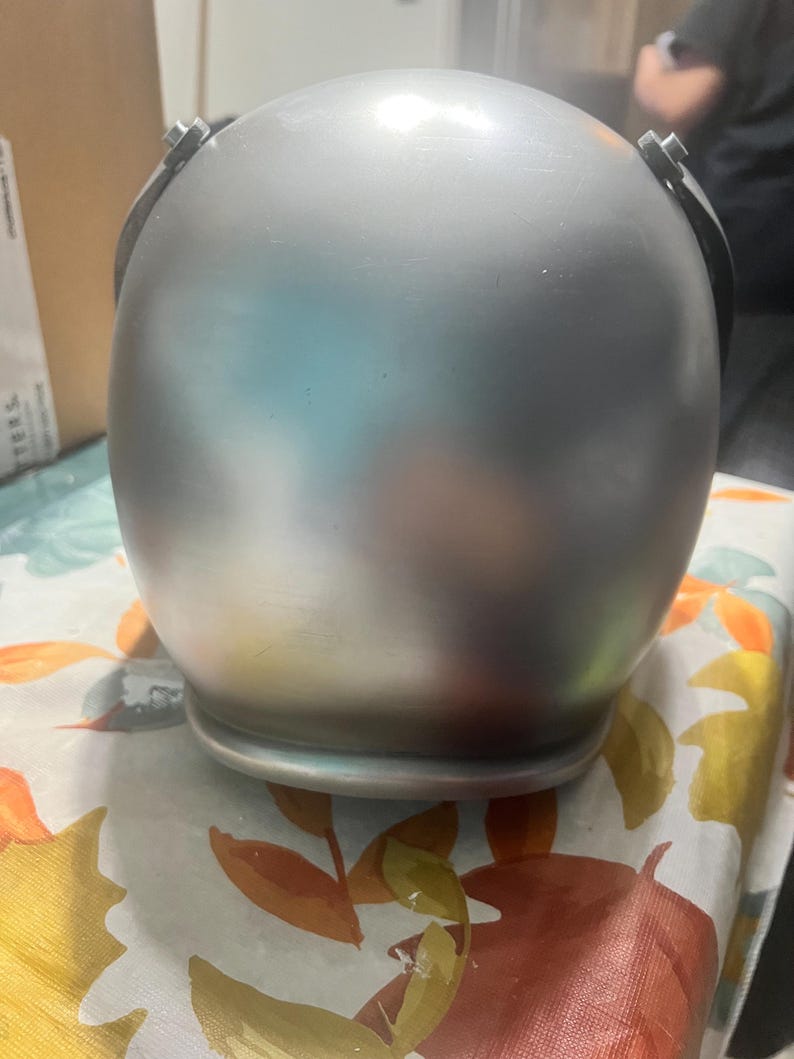 May include: A silver helmet with a smooth, shiny surface. The helmet has two metal clasps on the side.