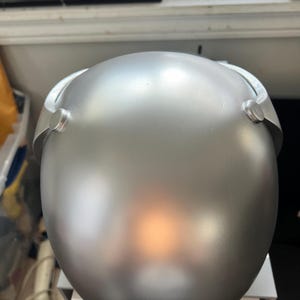 May include: A silver helmet with a smooth, matte finish. The helmet has two small, round protrusions on the sides.