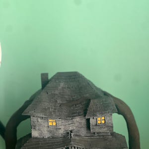 May include: A black and gray 3D printed model of a haunted house with glowing windows. The house has a gabled roof, a chimney, and a porch with railings. The house is sitting on a green surface.