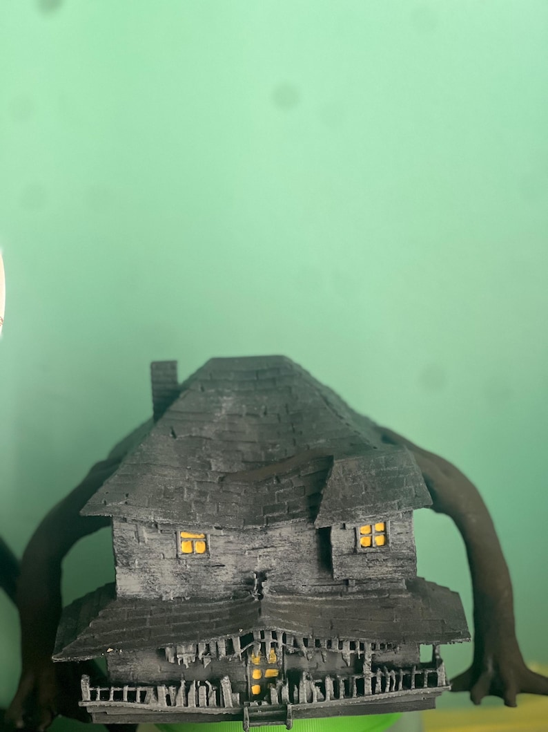 May include: A black and gray 3D printed model of a haunted house with glowing windows. The house has a gabled roof, a chimney, and a porch with railings. The house is sitting on a green surface.