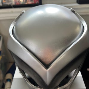 May include: A silver-colored helmet with a V-shaped design on the front. The helmet has a smooth, metallic finish.